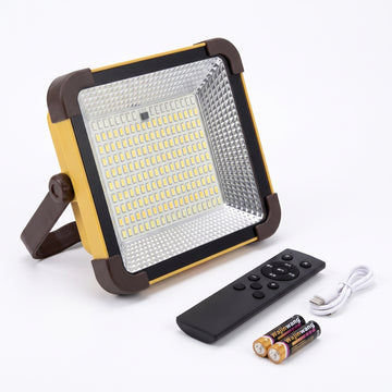 A square LED floodlight with a yellow and dark brown frame, shown with a black remote control, a white USB charging cable, and two batteries on a white background.